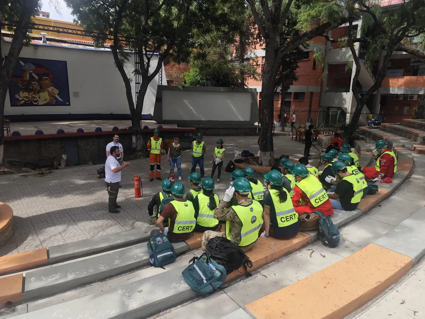 Grupo de voluntarios con cascos verdes y chalecos CERT sentados en el anfiteatro escuchan al instructor antes del simulacro