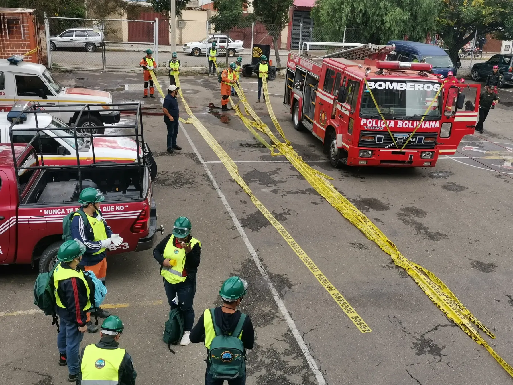 Vista elevada del simulacro: brigadistas tienden cintas de seguridad amarillas junto al carro bombero en el patio del cuartel