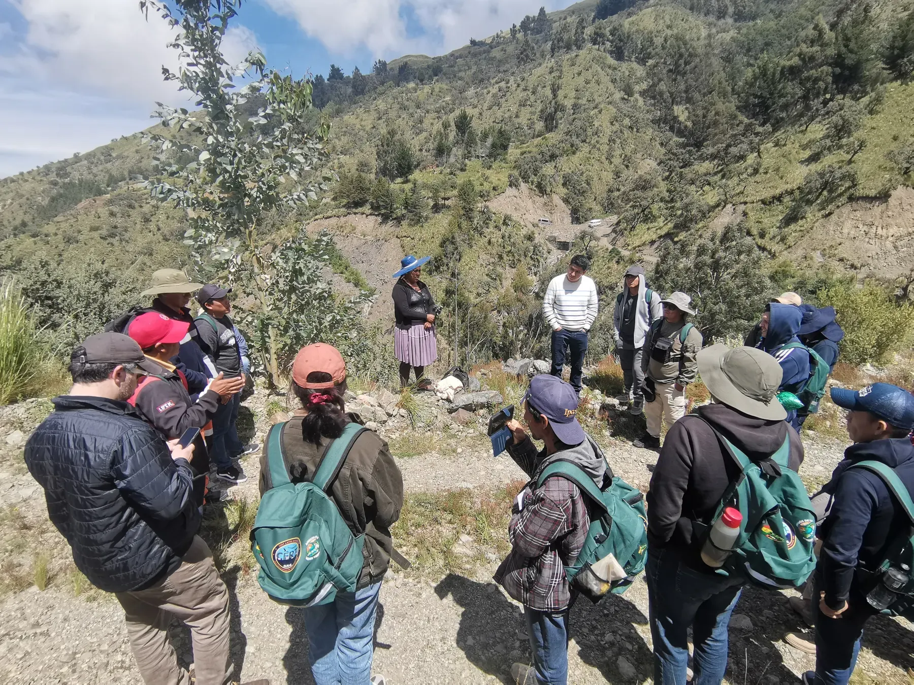 Círculo de brigadistas con mochilas verdes CERT escucha a la comunidad sobre la cuenca, con el valle andino de fondo